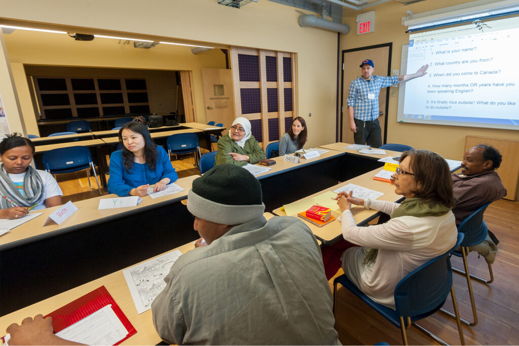 A group of students learning English together, sitting around tables as the instructor points at a list of questions projected on a large screen.
