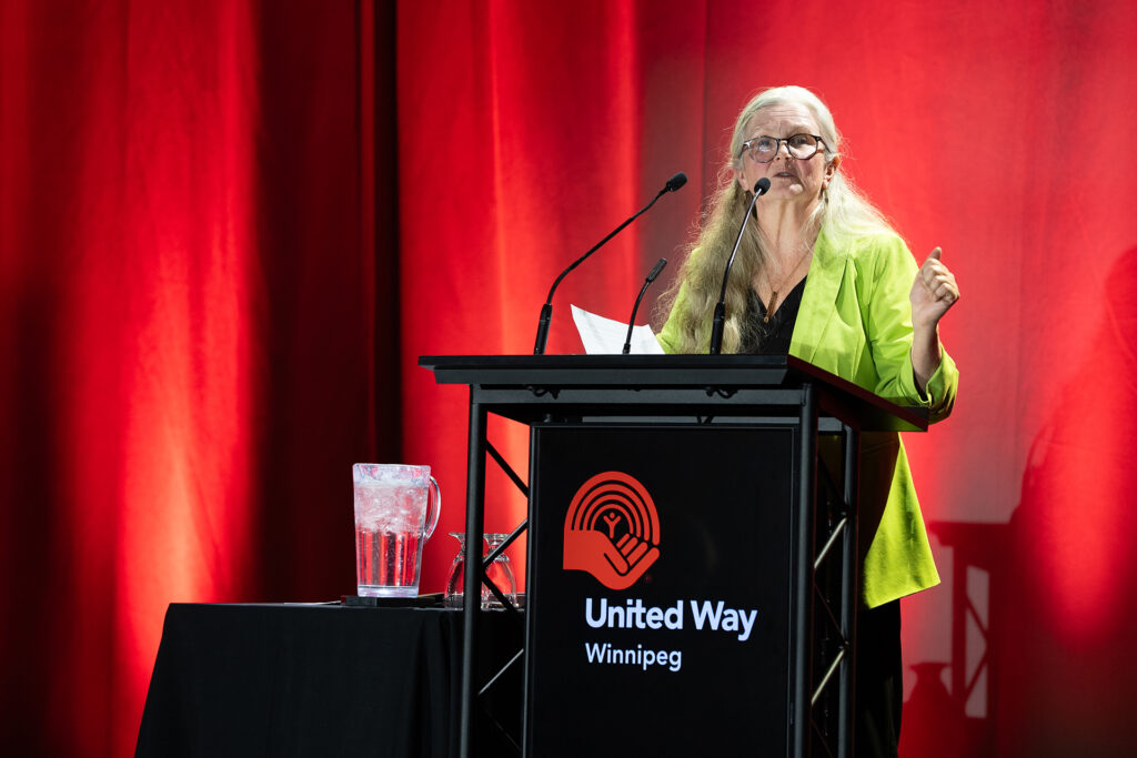 Cheryl sharing her story from a stage the RBC Convention Centre, a red United Way Winnipeg backdrop behind her.