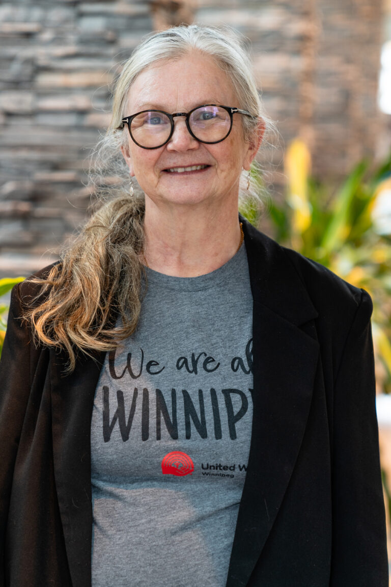 Cheryl smiles brightly while wearing a grey t-shirt that says "We Are All Winnipeg."