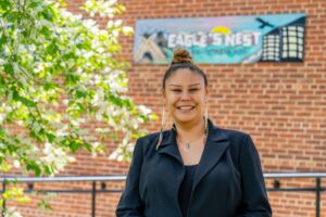 Desiree wears a black blazer and beautiful beaded earrings, while standing next to a blossoming cherry tree, with a painted sign for Eagle's Nest behind her.