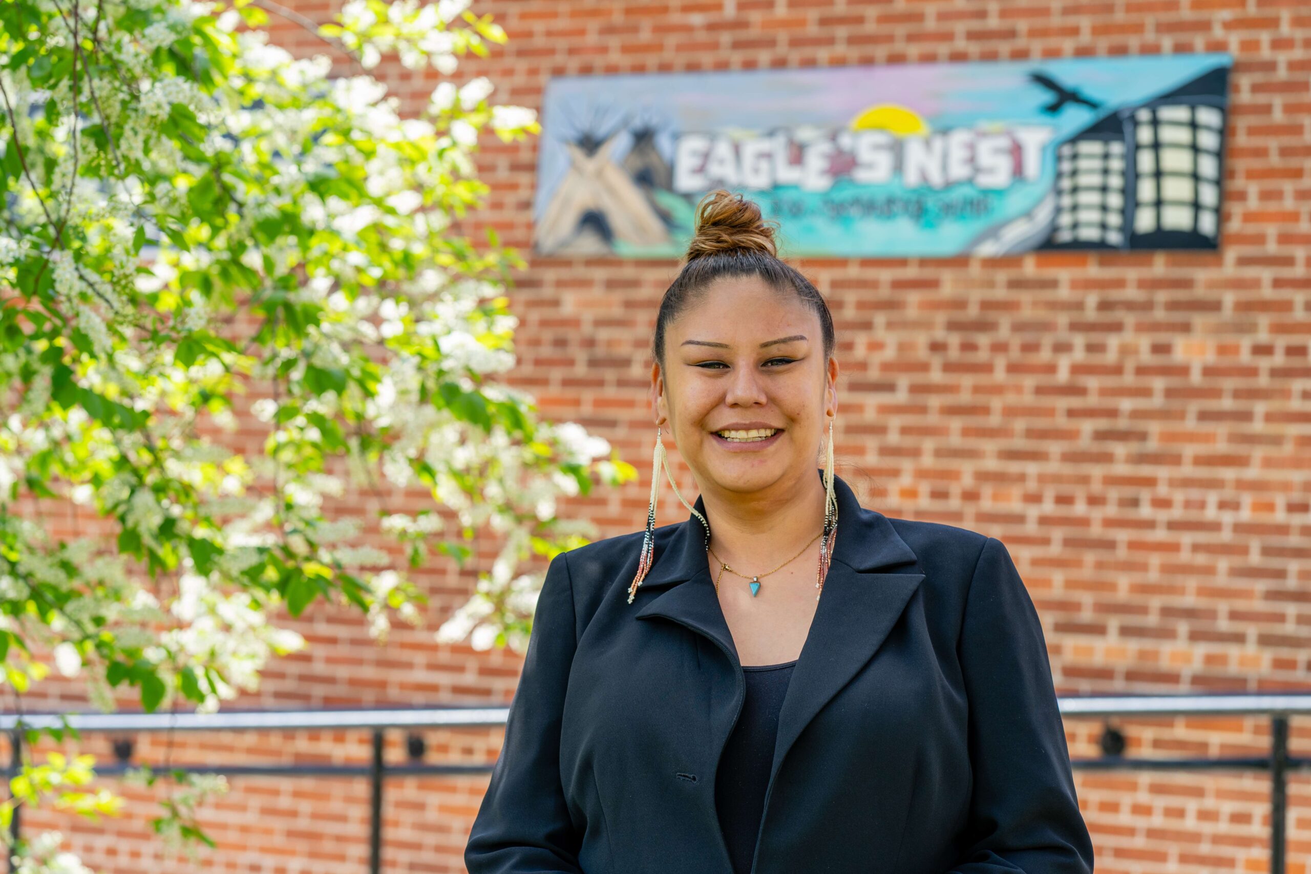 Desiree wears a black blazer and beautiful beaded earrings, while standing next to a blossoming cherry tree, with a painted sign for Eagle's Nest behind her.